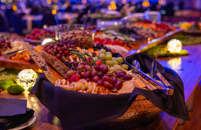 A long-serving table with food eloquently displayed like the main focus of a cedar basket with assorted bread, grapes, cheeses, and crackers. 
