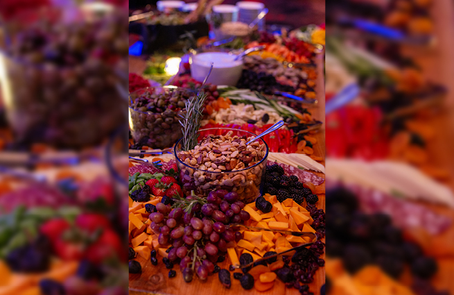 Food is displayed on a serving table. Assorted nuts in a glass bowl with a sprig of rosemary are surrounded by olives, strawberries, blackberries, and cheese.  