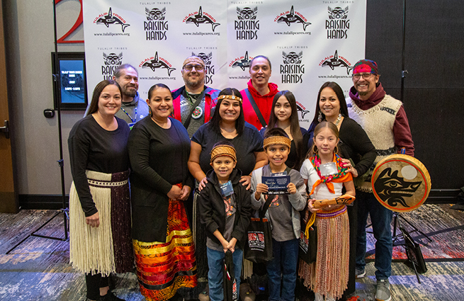 At the 2023 Raising Hands Event, a group of people with children in traditional Native American dress, pose for the camera. 