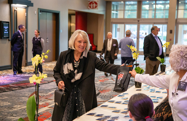 A woman arriving at the 2023 Raising Hands event being handed her gift bag at the reception table. People in the background. 