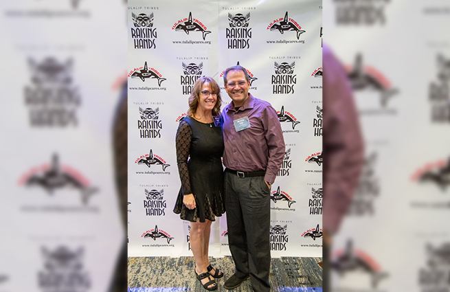 Couple posing for the camera in front the 2023 Raising Hands backdrop with their logo and the Tulalip Tribes whale logo displayed. 