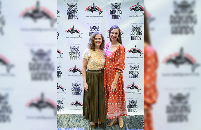 Two women standing together in front of the Raising Hands 2023 event backdrop, having their picture taken. They are smiling and happy. 