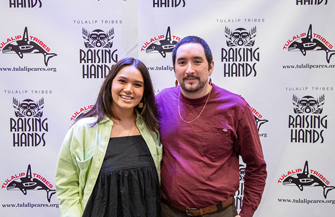 A couple having their picture taken in front of the 2023 Raising Hands backdrop banner. 