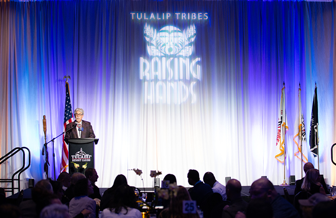 A man at the podium on the Tulalip Tribes Raising Hands stage, talking to the audience.