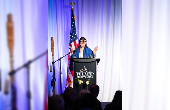 A woman with a traditional native headband and shawl, standing behind the podium for the 2023 Raising Hands event with an American flag behind her. Her hands are raised giving thanks. 