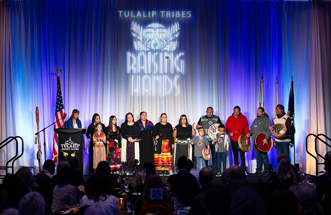 A group of adults and children stand on the 2023 Raising Hands stage, holding traditional hand-crafted drums, and many dressed in Native American attire. 