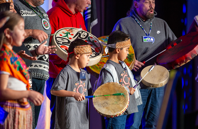 Two young Native American boys on the Raising Hands stage, wearing traditional Native American attire, playing deerskin drums.