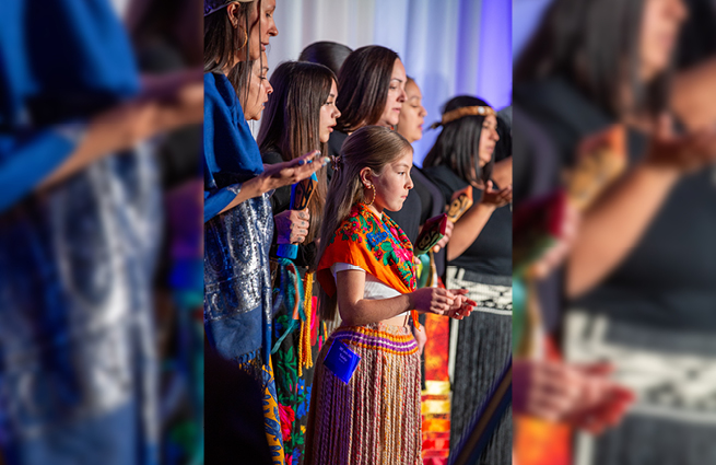 A young girl stands on stage with a group, wearing a beautiful, bright orange Native American outfit with a cedar woven skirt. 
