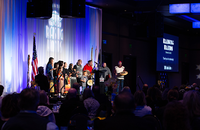 View from the audience of a group of adults and children of the tribal community, standing on stage, drumming deerskin drums, dressed in traditional attire. 