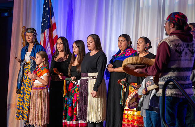 A group of beautiful tribal women and a young girl are singing on stage while being accompanied by tribal drummers. 