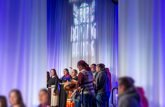 A group of adults and children standing on the Raising Hands stage in traditional Tulalip attire, singing and drumming handmade deerskin drums. 