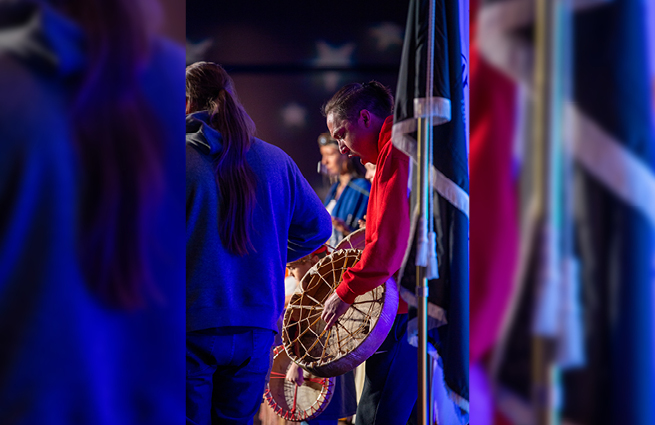 A man on the Raising Hands stage, standing by the flags, singing and drumming a Tulalip song of thanks. 