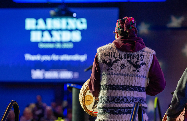 A view of a man from behind, wearing a handcraft wool vest with Williams stitched, drumming his beautiful handcrafted deerskin drum. 