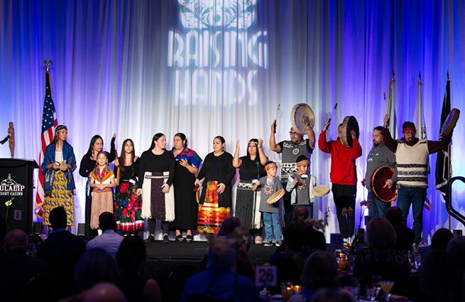 A group of adults and children of the Tulalip Tribes community, gathered on stage, singing and drumming, with raised hands in thanks.  