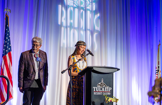 View a woman addressing the Raising Hands audience from behind the podium, concerning Tulalip Cares' charity work. Mel Sheldon stands off to the side. 