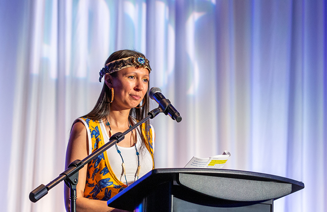 A woman in traditional tribal attire and a handcrafted cedar headband stands behind the podium addressing attendees. 