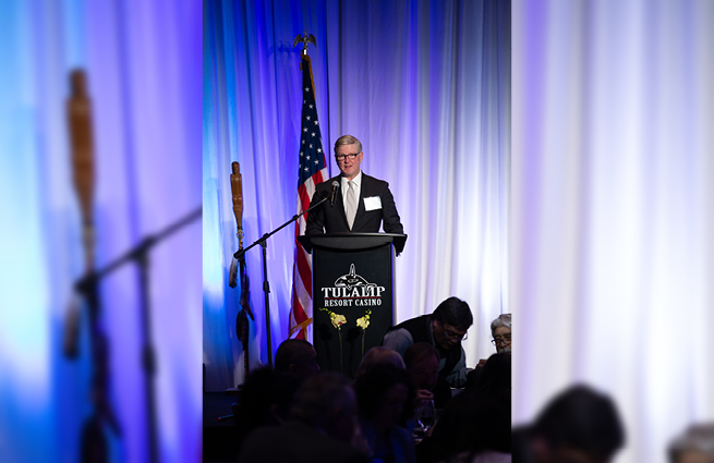 A man stands behind the podium in the banquet room at Tulalip Casino Resort speaking to attendees at the 2023 Raising Hands event. 