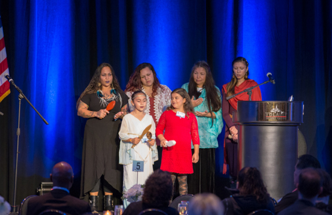 Tulalip Tribes’ 2018 Raising Hands photo of a ceremony with traditional singing, including drumming, raising hands from the group’s right.