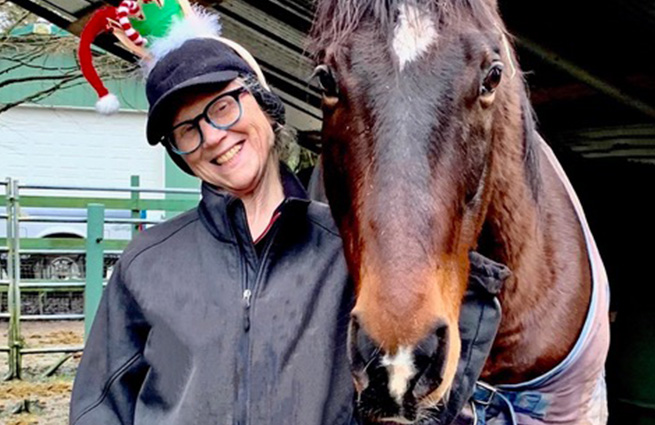 An equine rescue volunteer, wearing an elf hat, stands next to a rescued bay horse. 