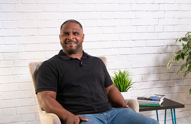 A blind man, smiling and sitting in a white chair with a small stack of books beside him on a small table with a plant. 