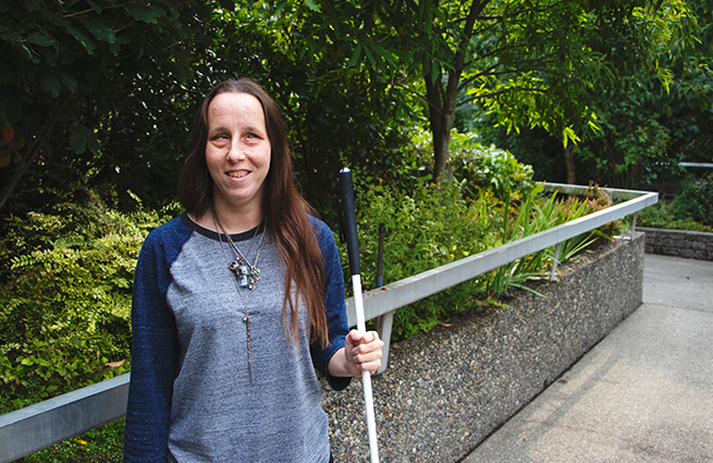 Blind young woman, with her cane, standing on a concrete walkway along a contained garden area behind a concrete wall. 