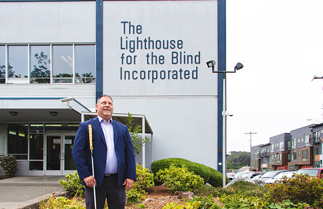 Blind man with blue blazer and cane, standing in front of The Lighthouse for the Blind Incorporated building.