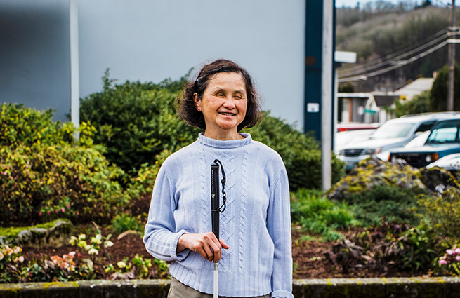 Blind woman in a light blue sweater, with her cane, standing in front of The Lighthouse for the Blind Corporation office building. 