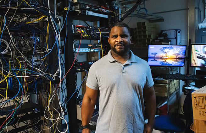 A man standing in a room of technical equipment behind him and computer monitors next to him. 