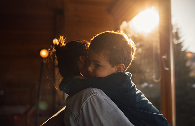 A mother holding a toddler boy in her arms on a porch with the sun setting. 