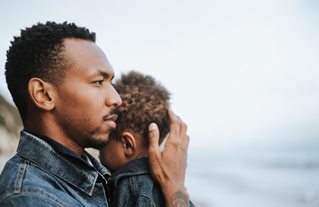 A young father, sweetly holding his toddler daughter with his hand behind her head, standing on the beach, looking out over the waves breaking. 