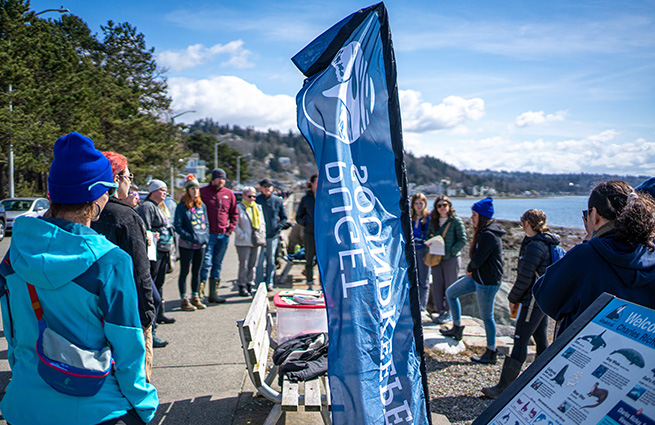 Puget Soundkeeper's volunteers at Charles Richey Sr. Viewpoint park gather around a parkbench on a sunny day at the water's edge. 