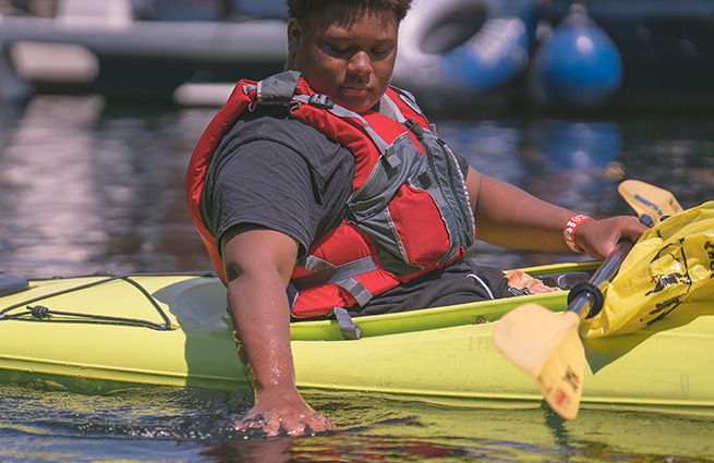 A young man floating on the water in an inflatable kayak, touching the top of the water with his hand.  