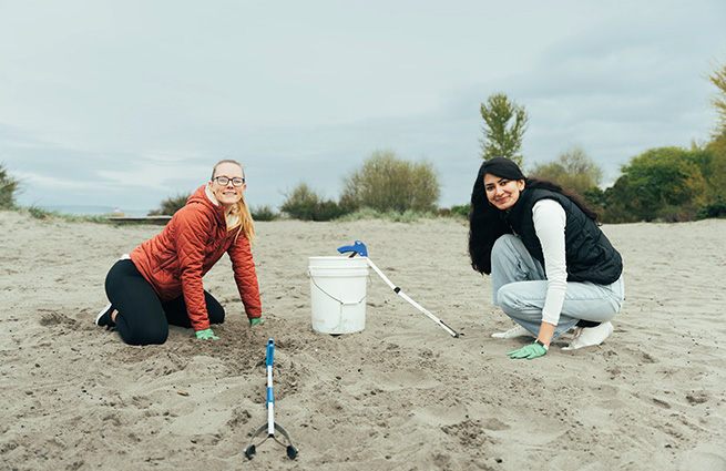 Under cloudy skies, two women crouch on a white sand beach, with a white 5-gallon bucket between them and grippers to clean up the beach.
