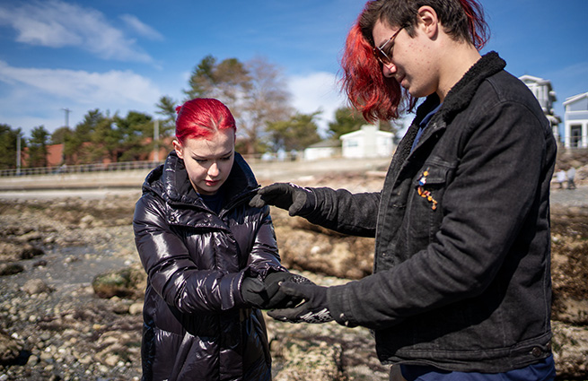 A young woman and a young man, standing on a rocky beach, observe a beach lifeform held in the young woman's hands. 