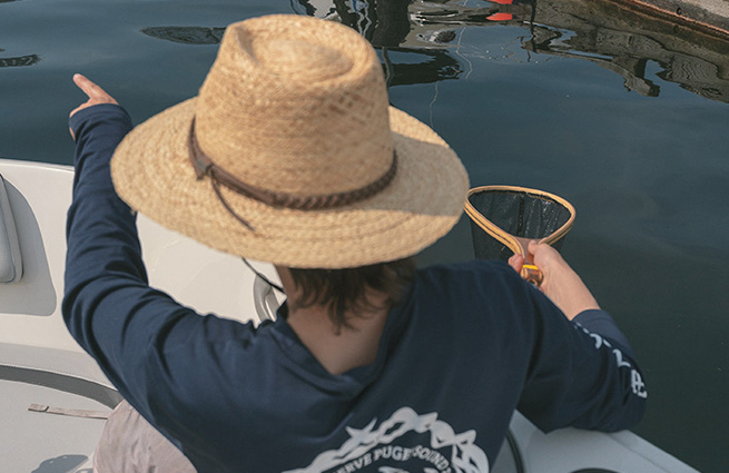 A person in a boat, wearing a Puget Soundkeeper long-sleeved shirt, and a big sun hat is cleaning litter out of the bay. 