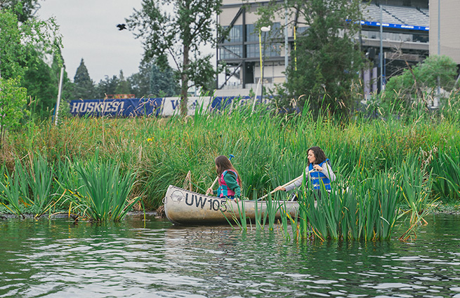 Two women in a metal canoe, along the shoreline with Huskies stadium in the background, tucked inside tall foliage, use nets to scoop and clear out debris. 