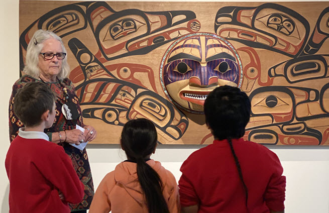 Students gathered around tribal wall art with an adult presenter describing the piece. 
